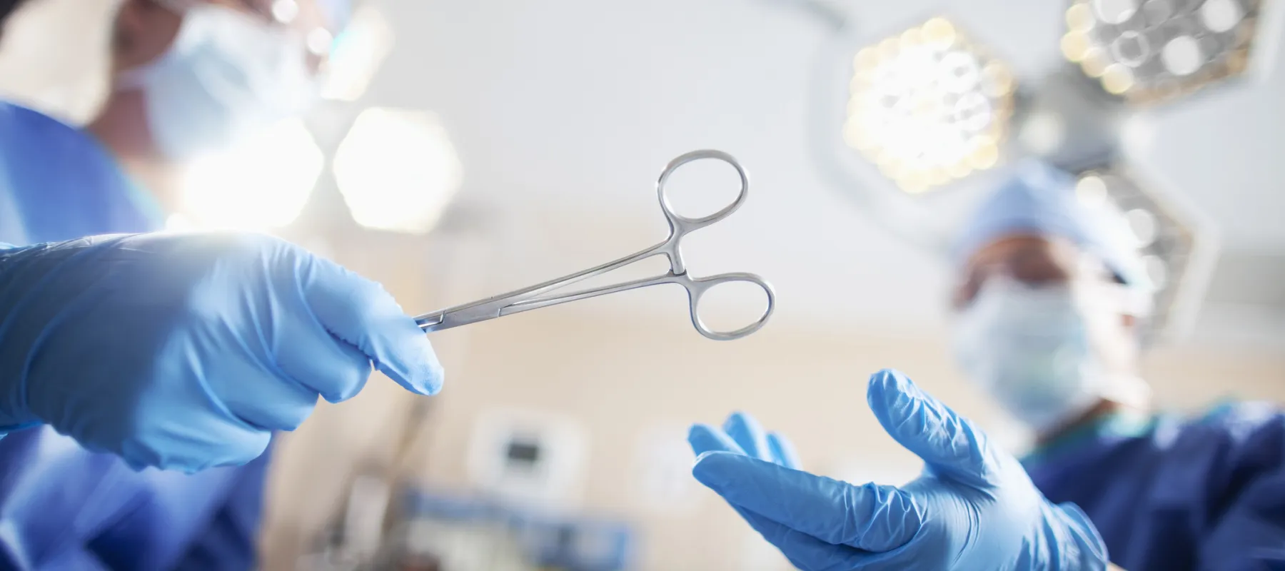 Surgeons wearing blue gloves and masks passing surgical forceps in an operating room under bright lights.