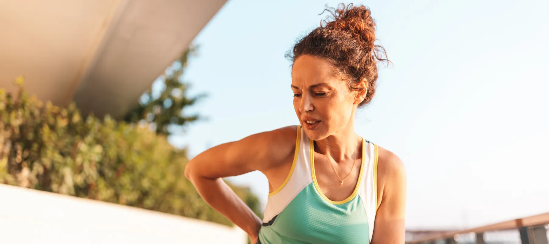 Woman in athletic wear holding her side in pain while running outdoors on a sunny day.