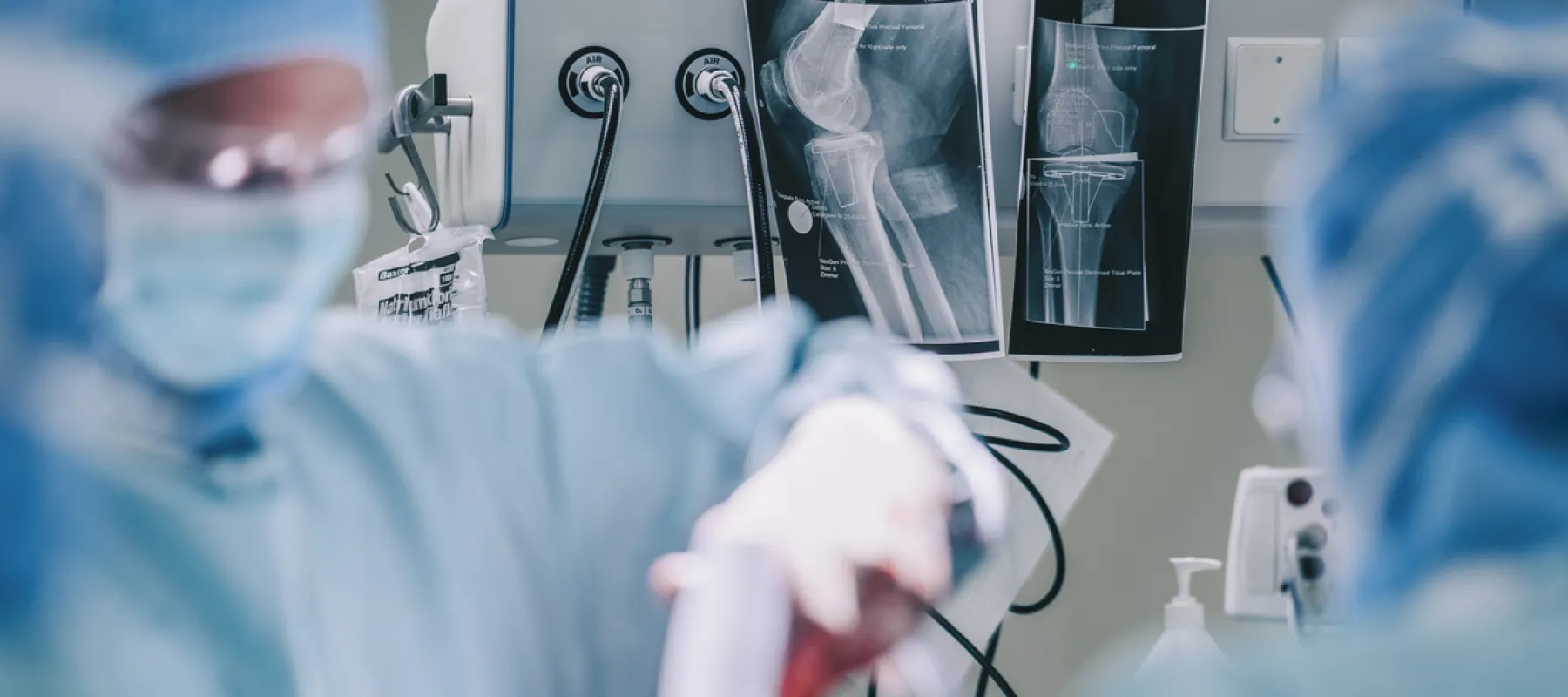 Surgeons in protective gear performing surgery with X-ray images of a knee joint displayed in the background