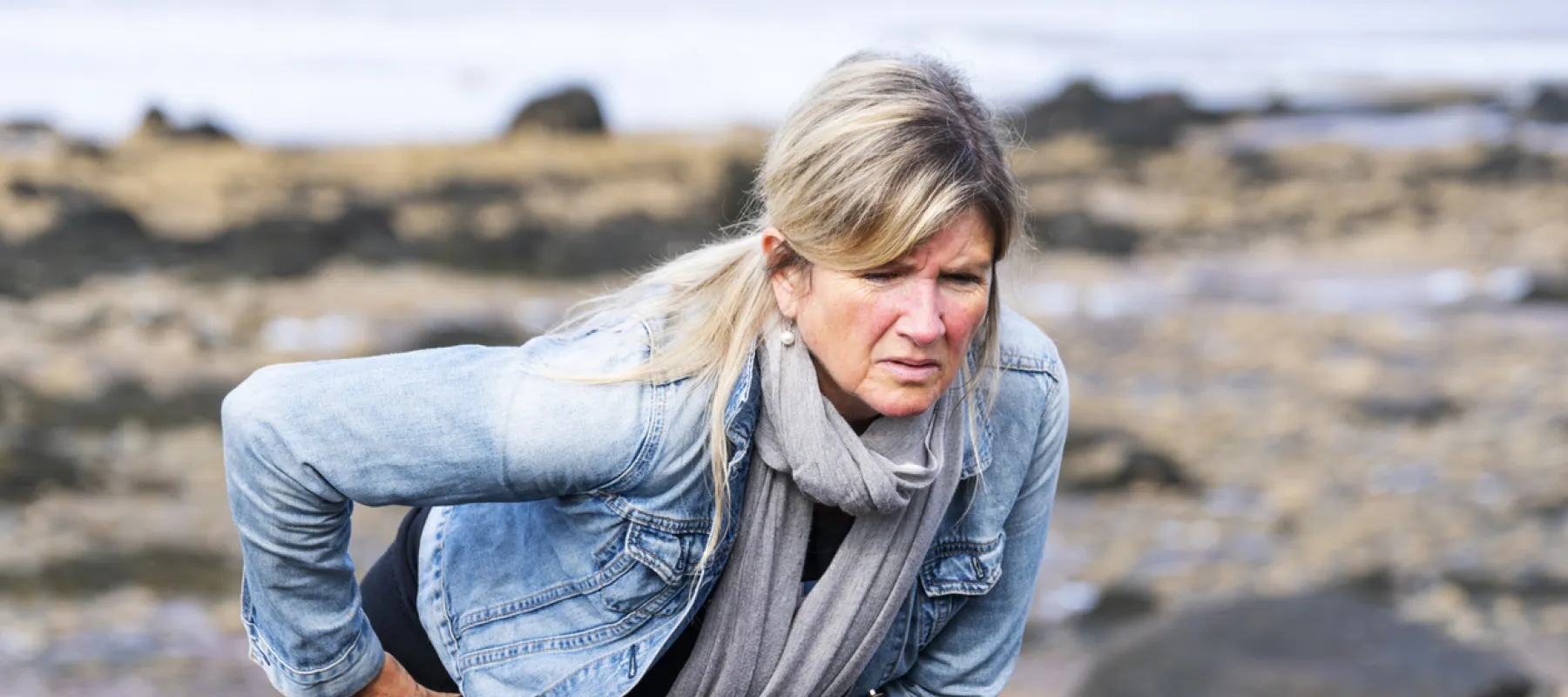 Mature woman in denim jacket and scarf holding hip in pain while standing on rocky beach shore.