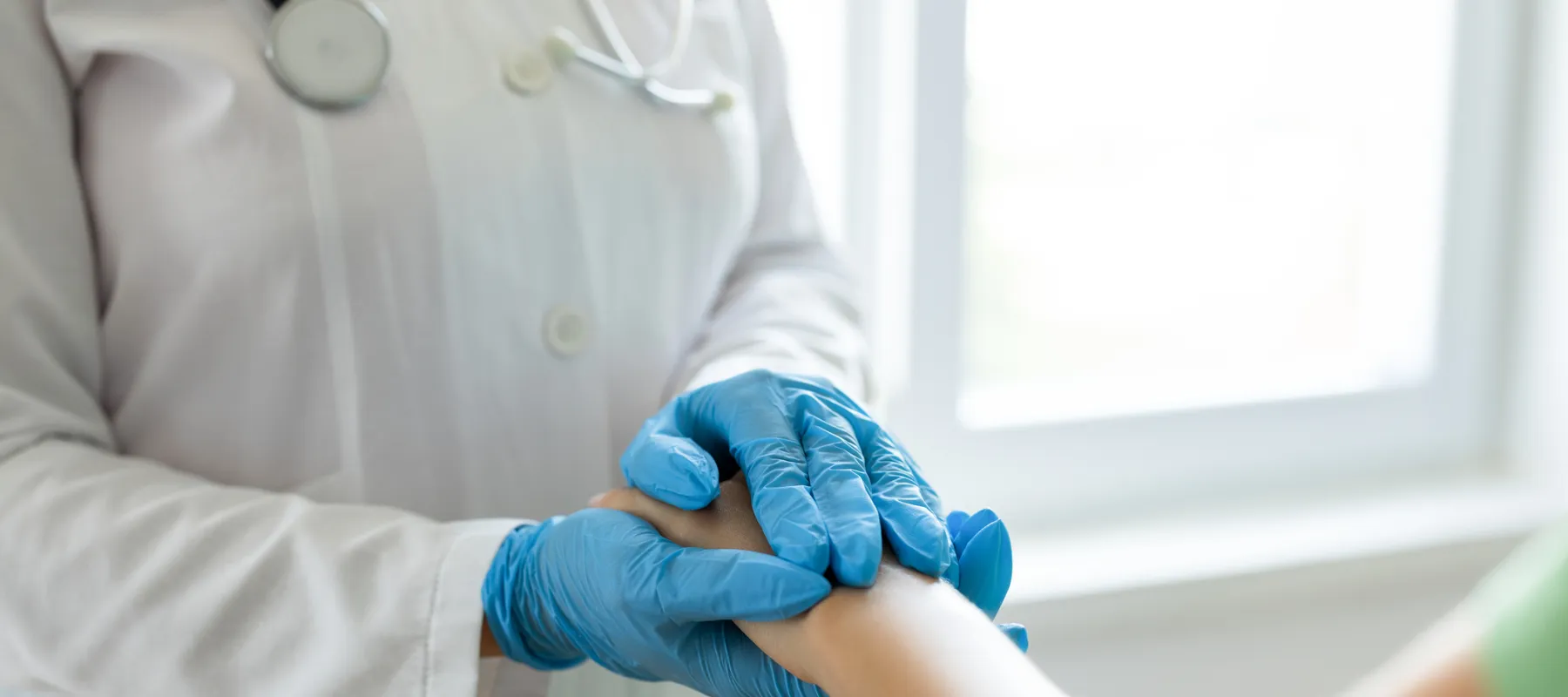 Doctor in white coat with stethoscope wearing blue gloves holding patient wrist in medical consultation.