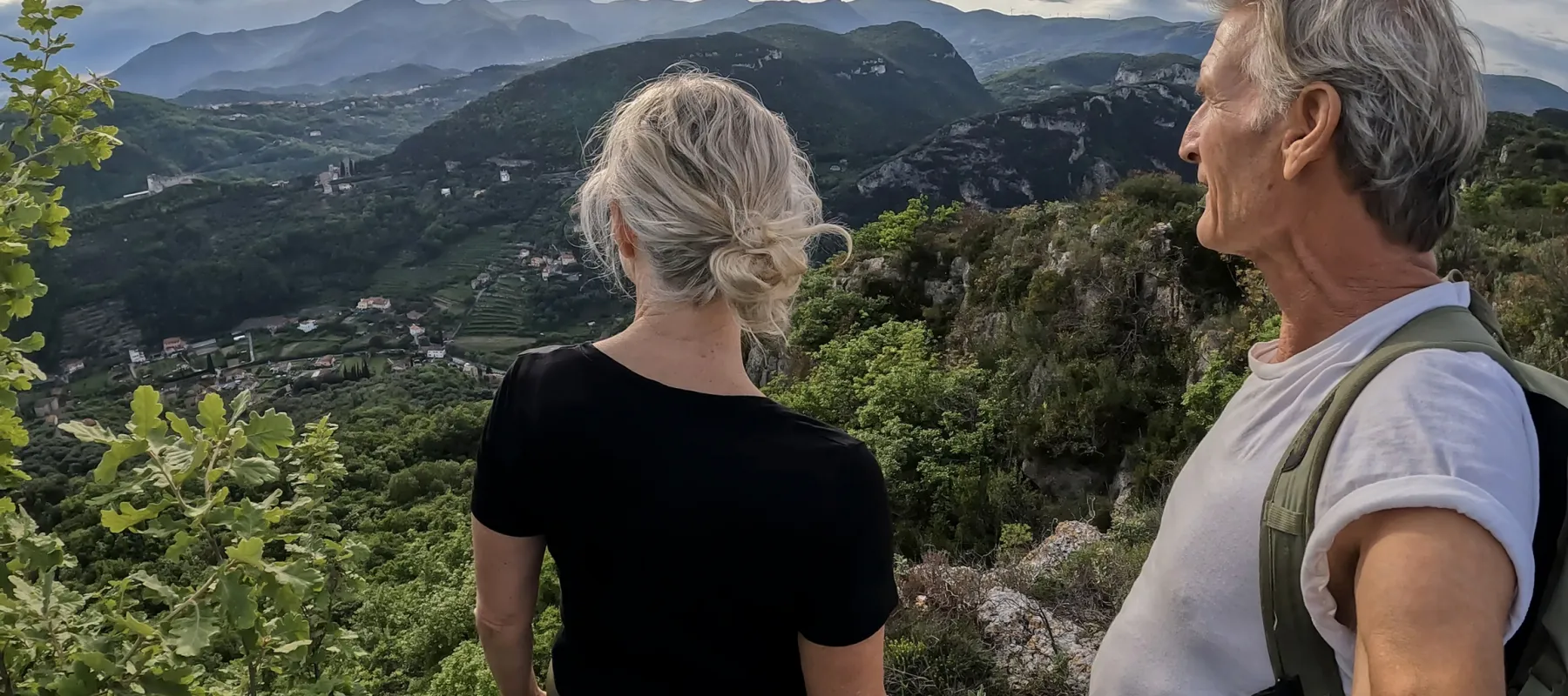 Couple hiking atop a mountain overlooking a lush green valley and distant mountains under cloudy sky.