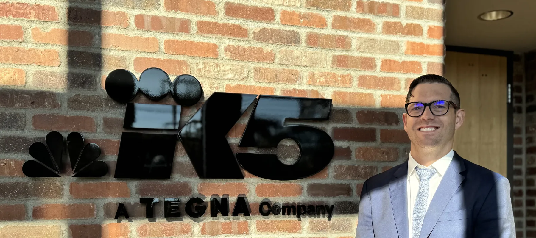 Man in blue suit standing next to K5 Tegna Company logo on brick wall in sunlight.