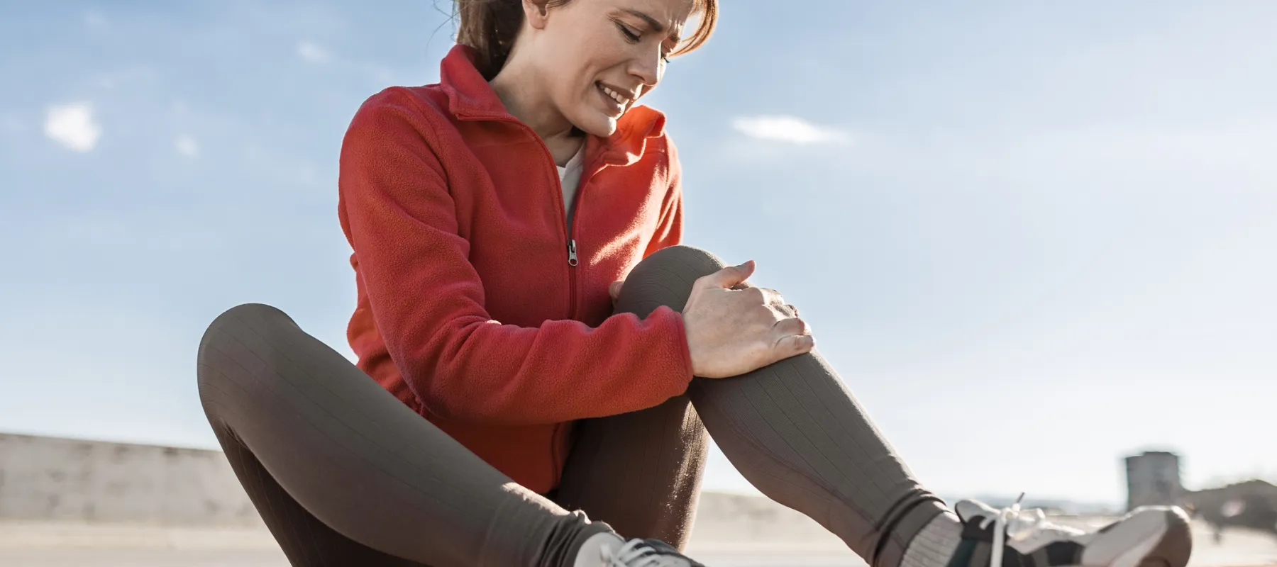 Young woman in red jacket sitting on track holding her painful knee outdoors under clear sky