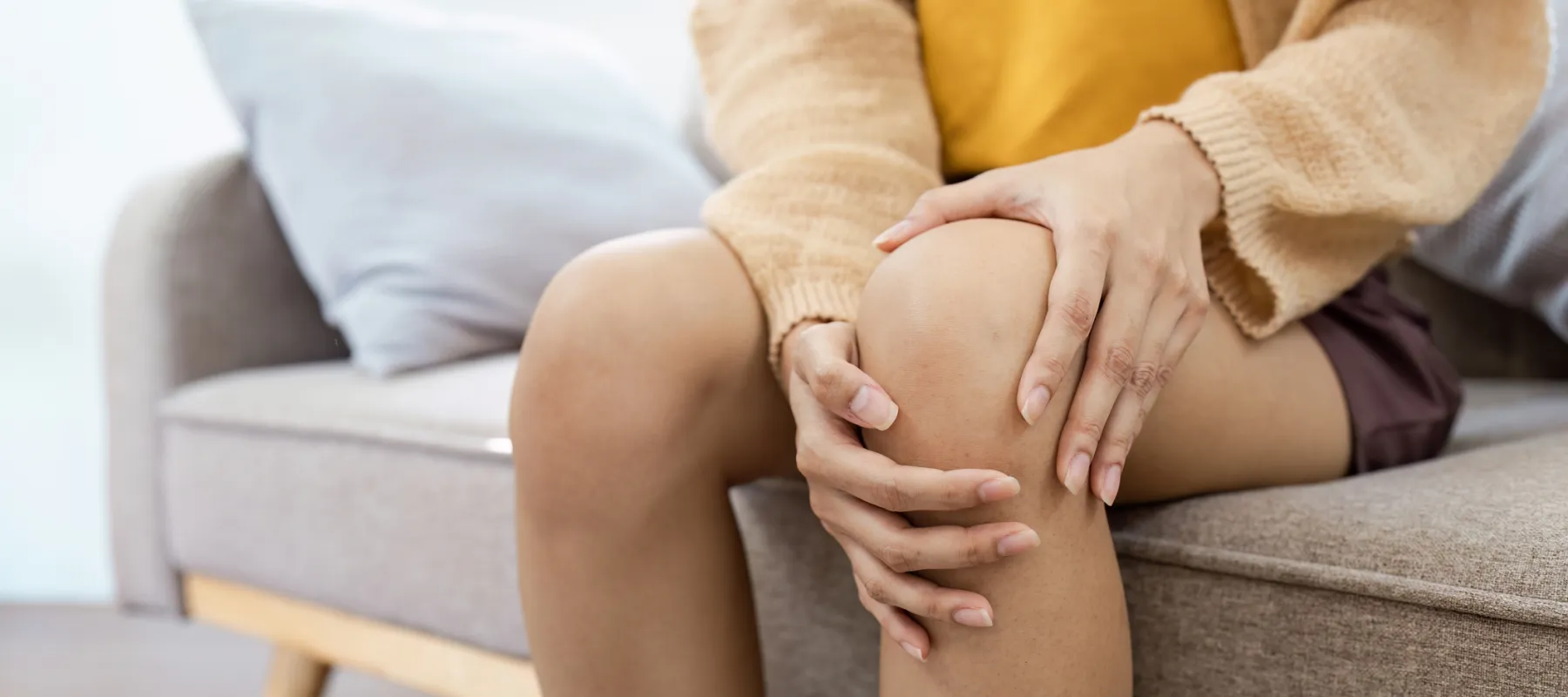Person sitting on a sofa holding their knee, indicating knee pain or discomfort indoors.
