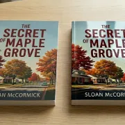 A matte laminated copy and a gloss laminated copy of The Secret of Maple Grove by Sloan McCormick on a wooden table with autumn trees on the cover.