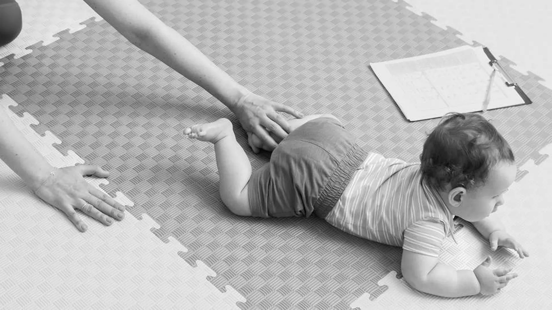 Baby crawling on foam play mat with an adult kneeling nearby and a clipboard on the floor