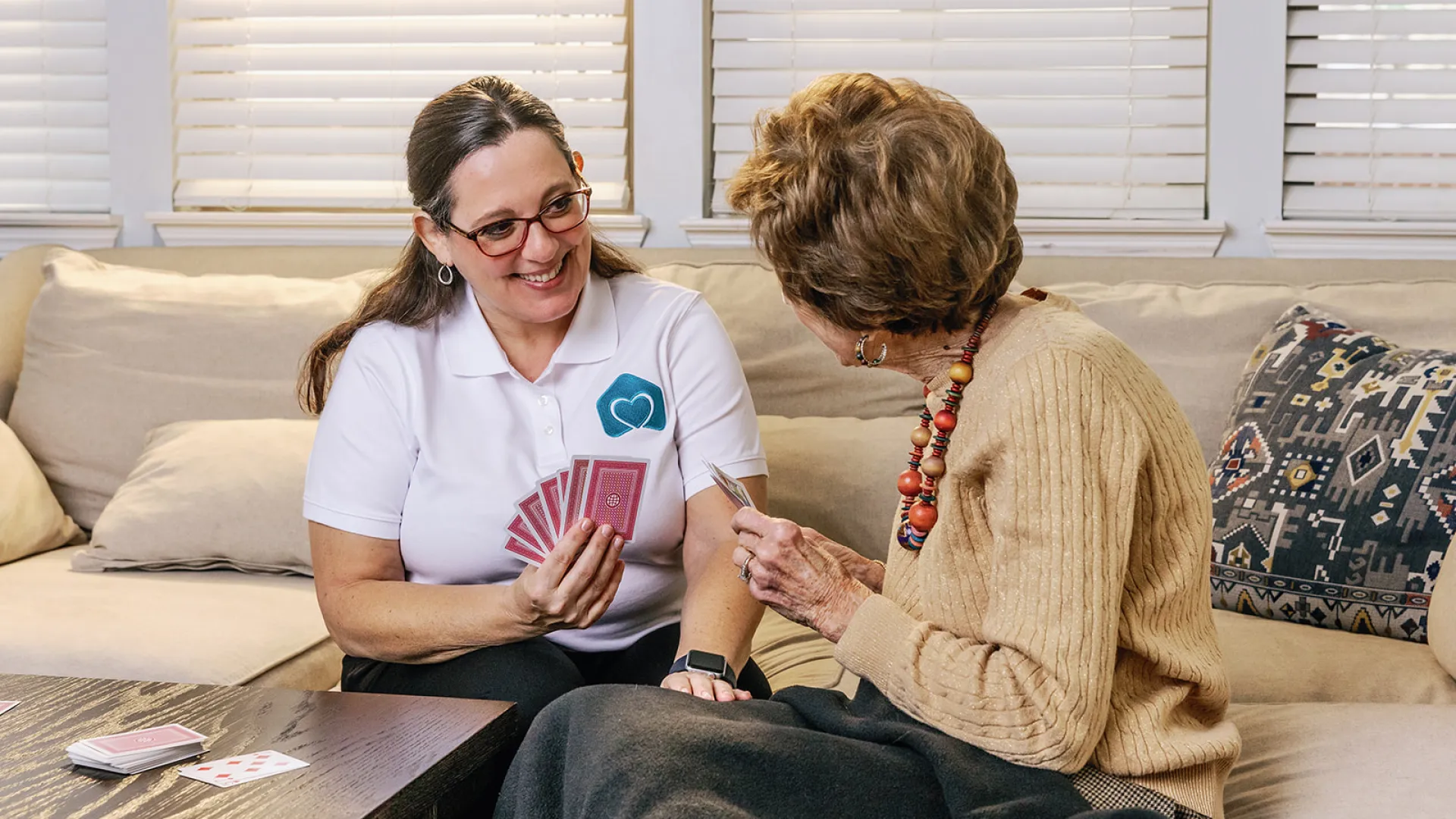 Caregiver playing cards and smiling with elderly woman on sofa in cozy living room setting