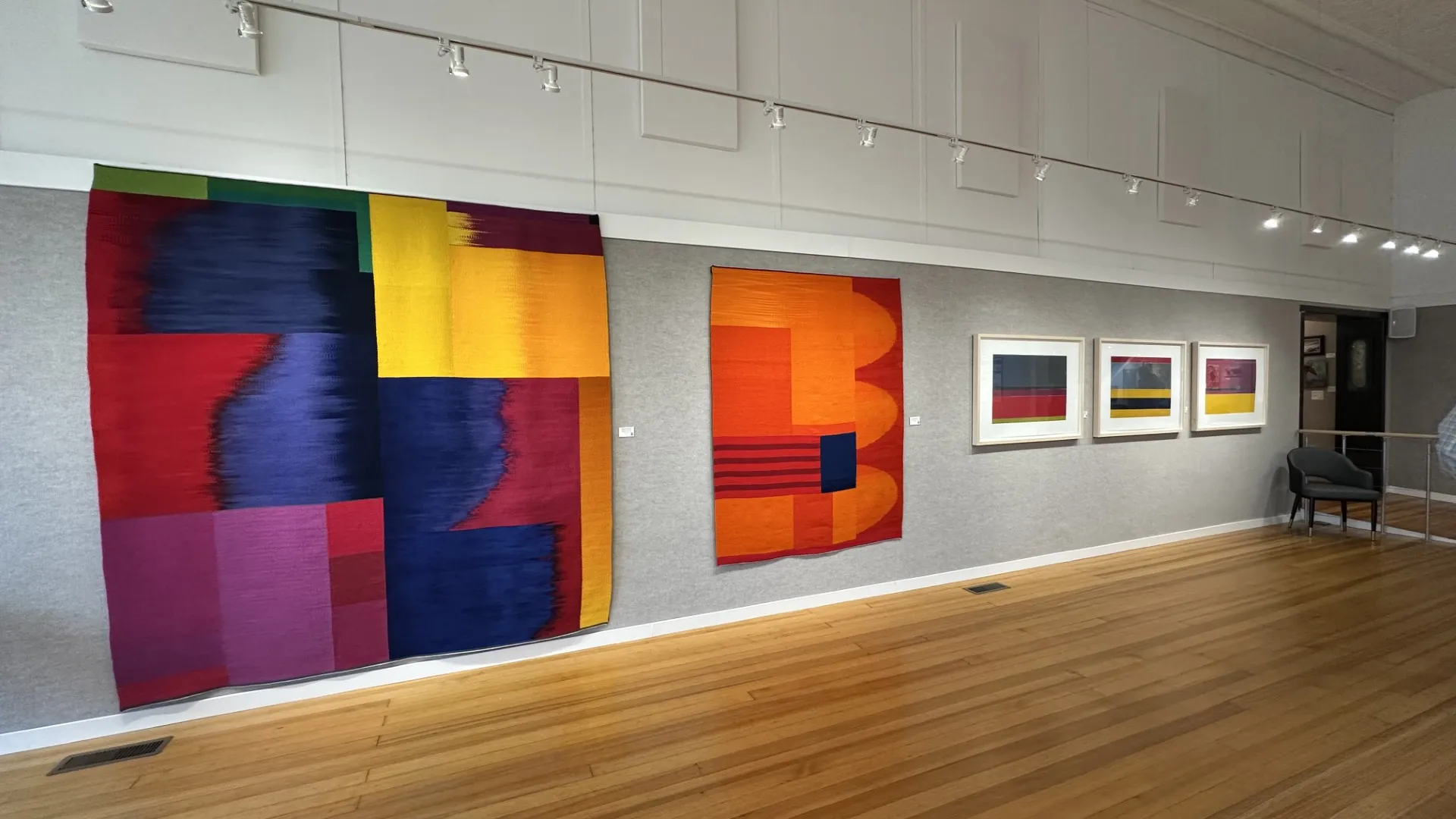 Colorful abstract textile artworks and framed prints displayed on gallery wall with wooden floor and track lighting.