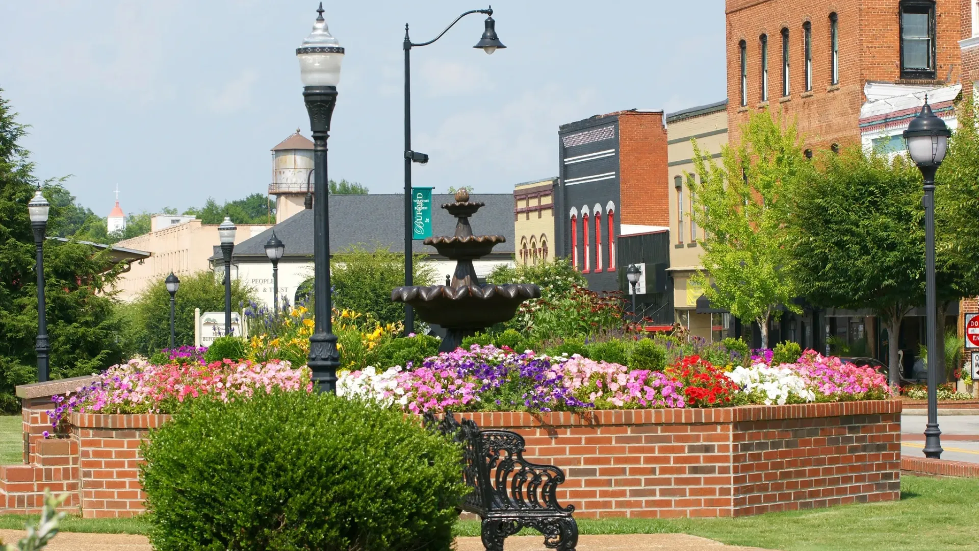 Colorful flower bed with fountain, iron bench, and historic brick buildings in a small town downtown park.