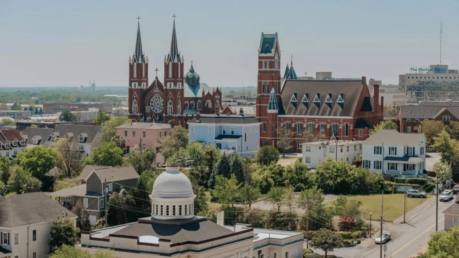 Aerial view of a historic town with churches, a courthouse with a dome, residential houses, and a hospital in the background.
