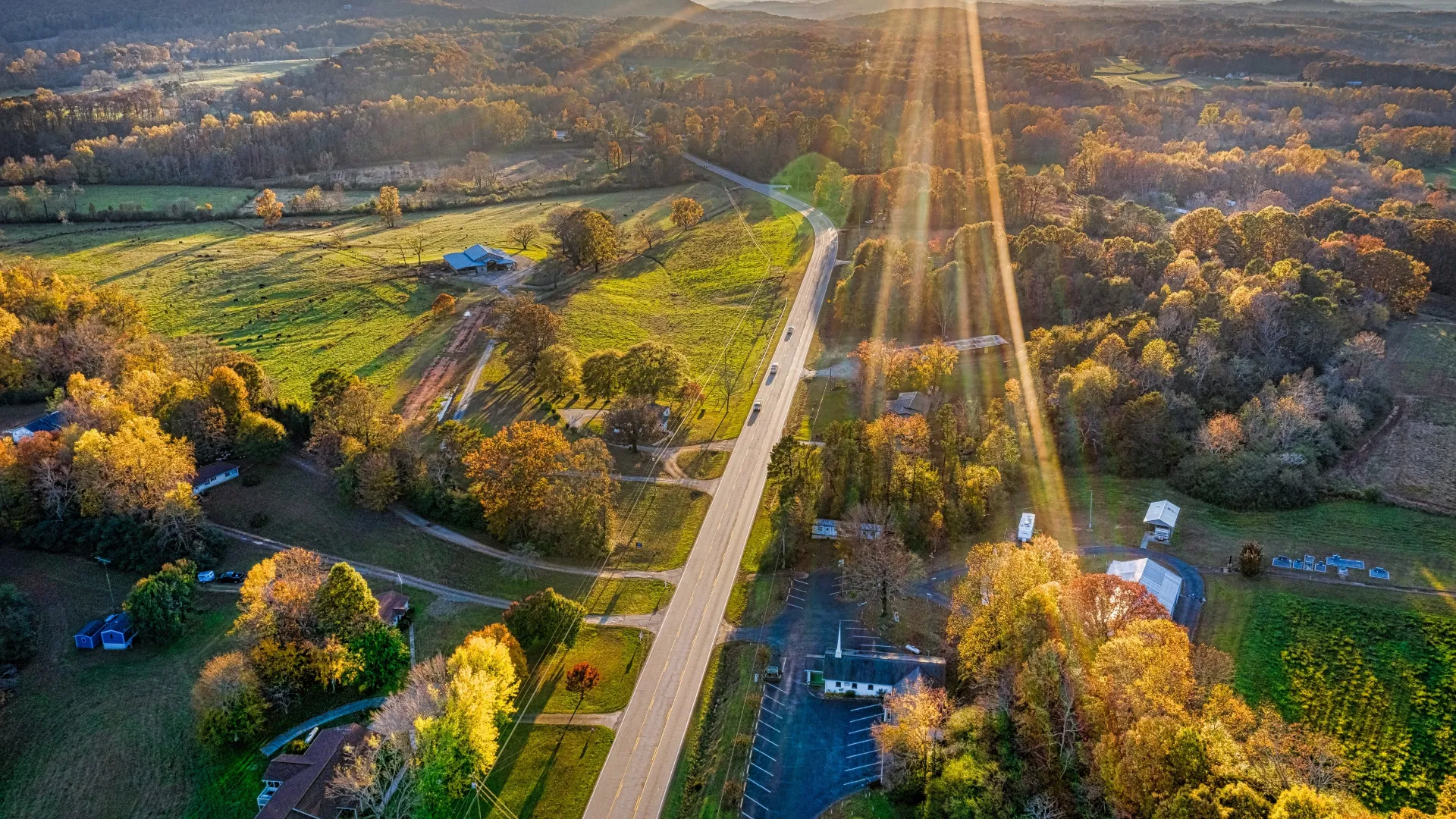 Sun rays over winding rural road surrounded by autumn trees and rolling hills at sunset.