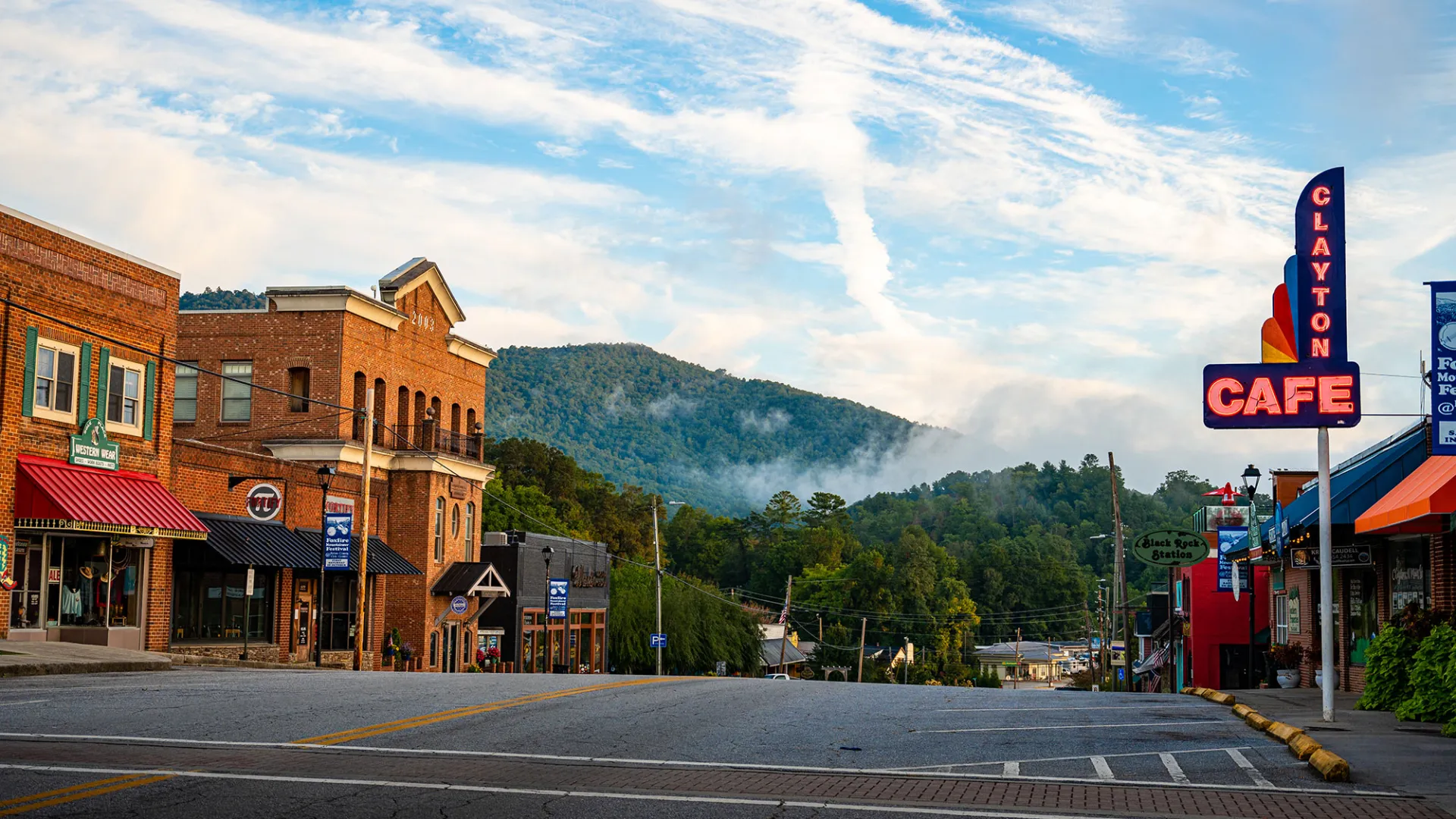 Historic small town street with brick buildings, Clayton Cafe sign, and forested mountains under a blue sky.