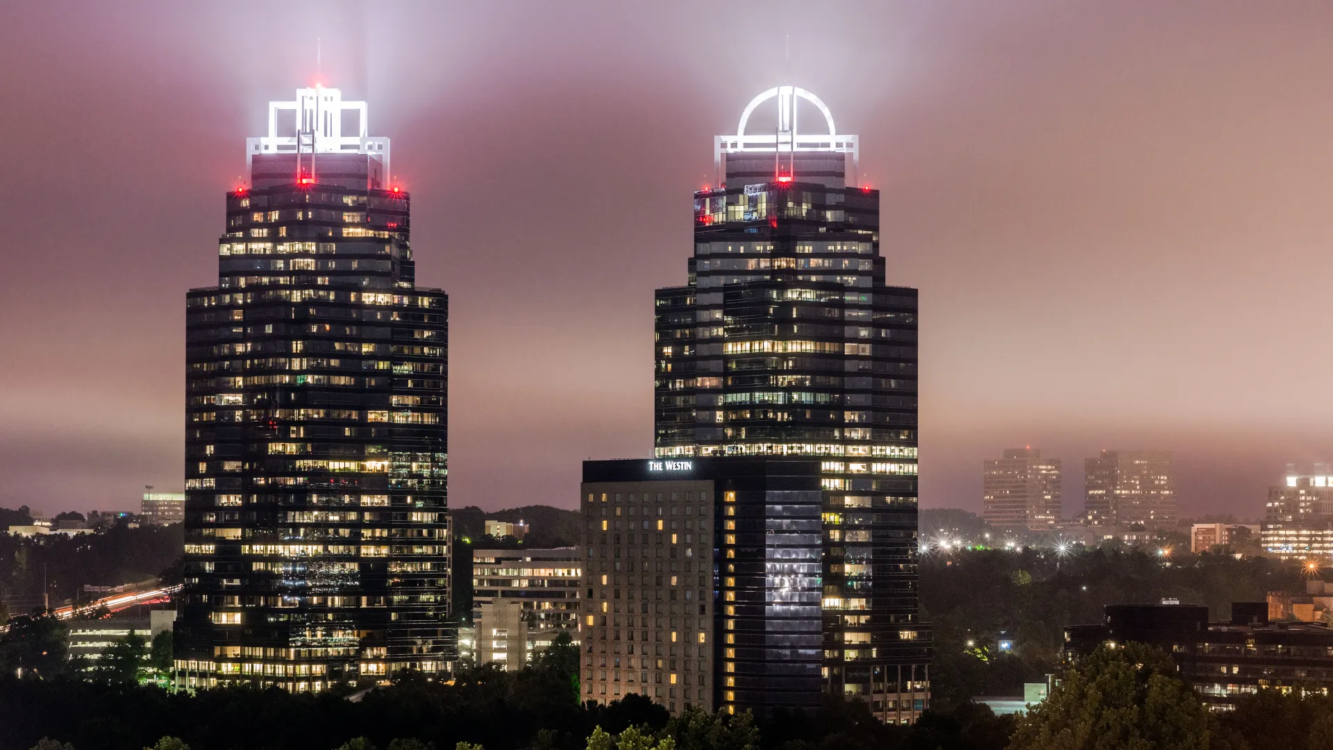 Night view of two illuminated skyscrapers with distinctive crown-shaped tops and surrounding cityscape in foggy weather
