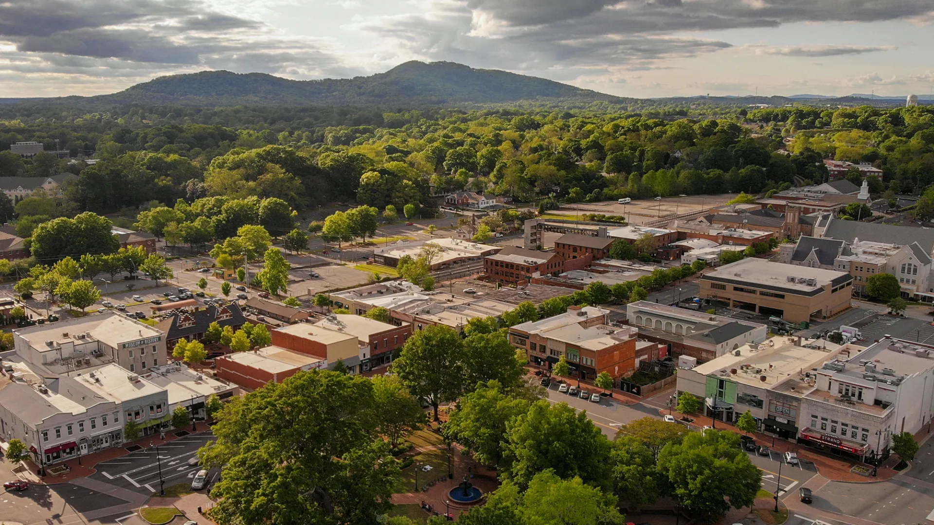 Aerial view of marietta with green trees, buildings, streets, and mountains under a cloudy sky at sunset.