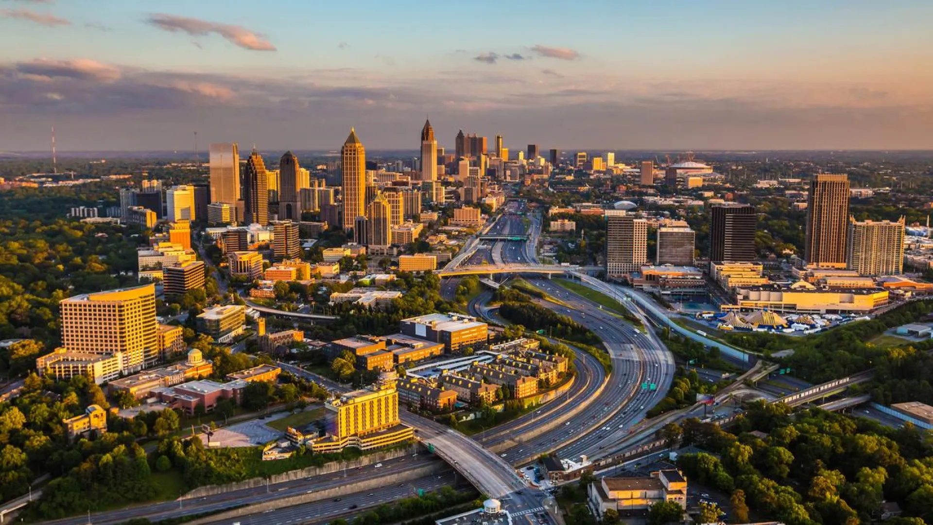 Aerial view of Atlanta city skyline with highways and green trees during sunset under a partly cloudy sky.