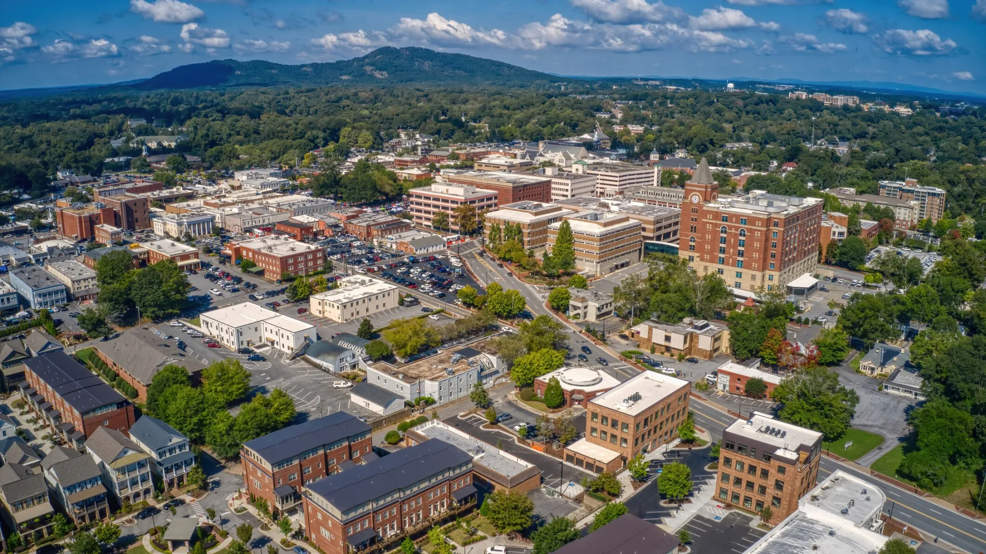 Aerial view of a small city with residential and commercial buildings surrounded by green hills under a blue sky.