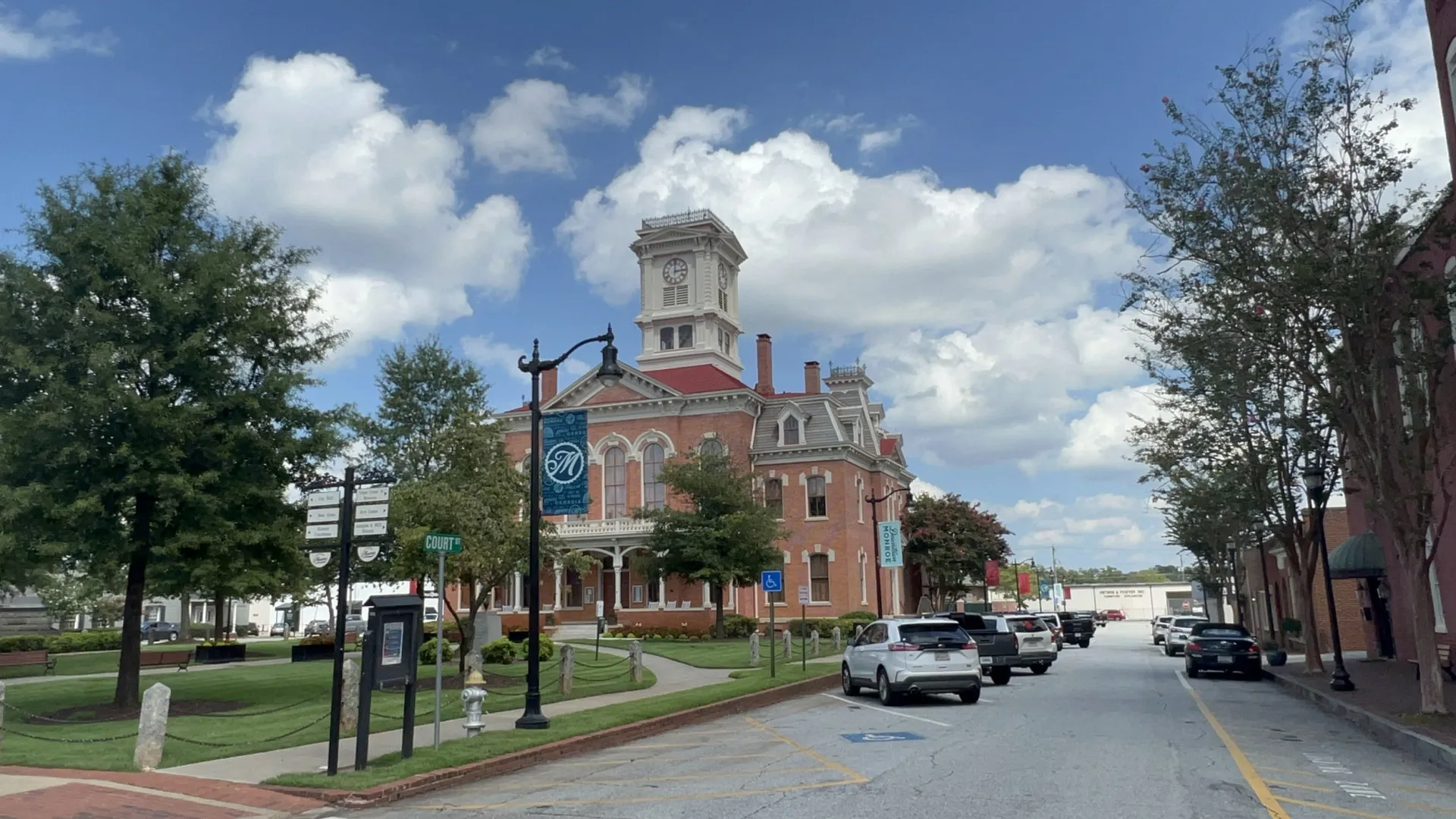 Historic brick courthouse with clock tower on a tree-lined street under a blue sky with clouds Monroe GA
