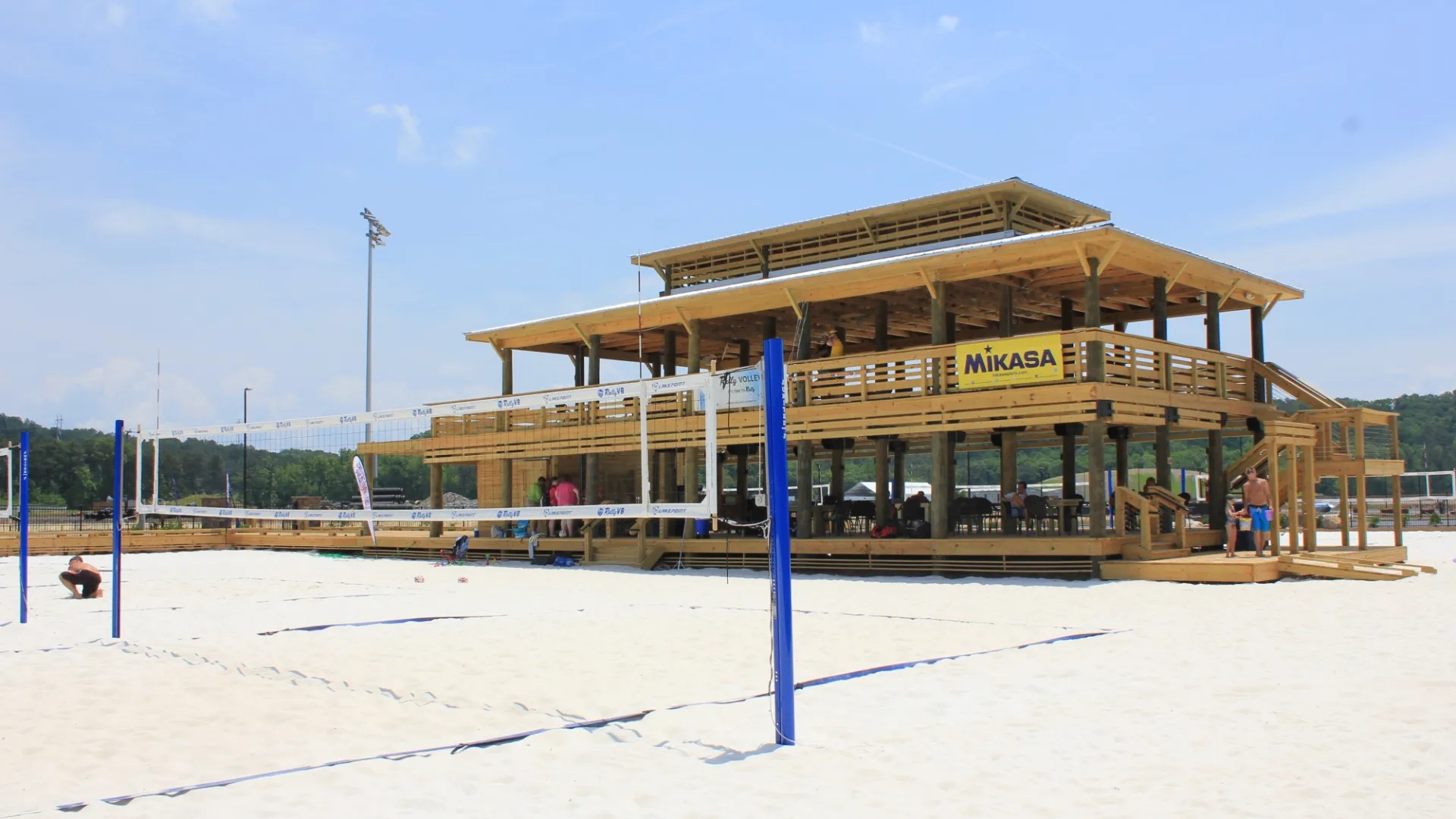 Beach volleyball courts with nets and a large wooden pavilion under a clear blue sky on sandy ground.
