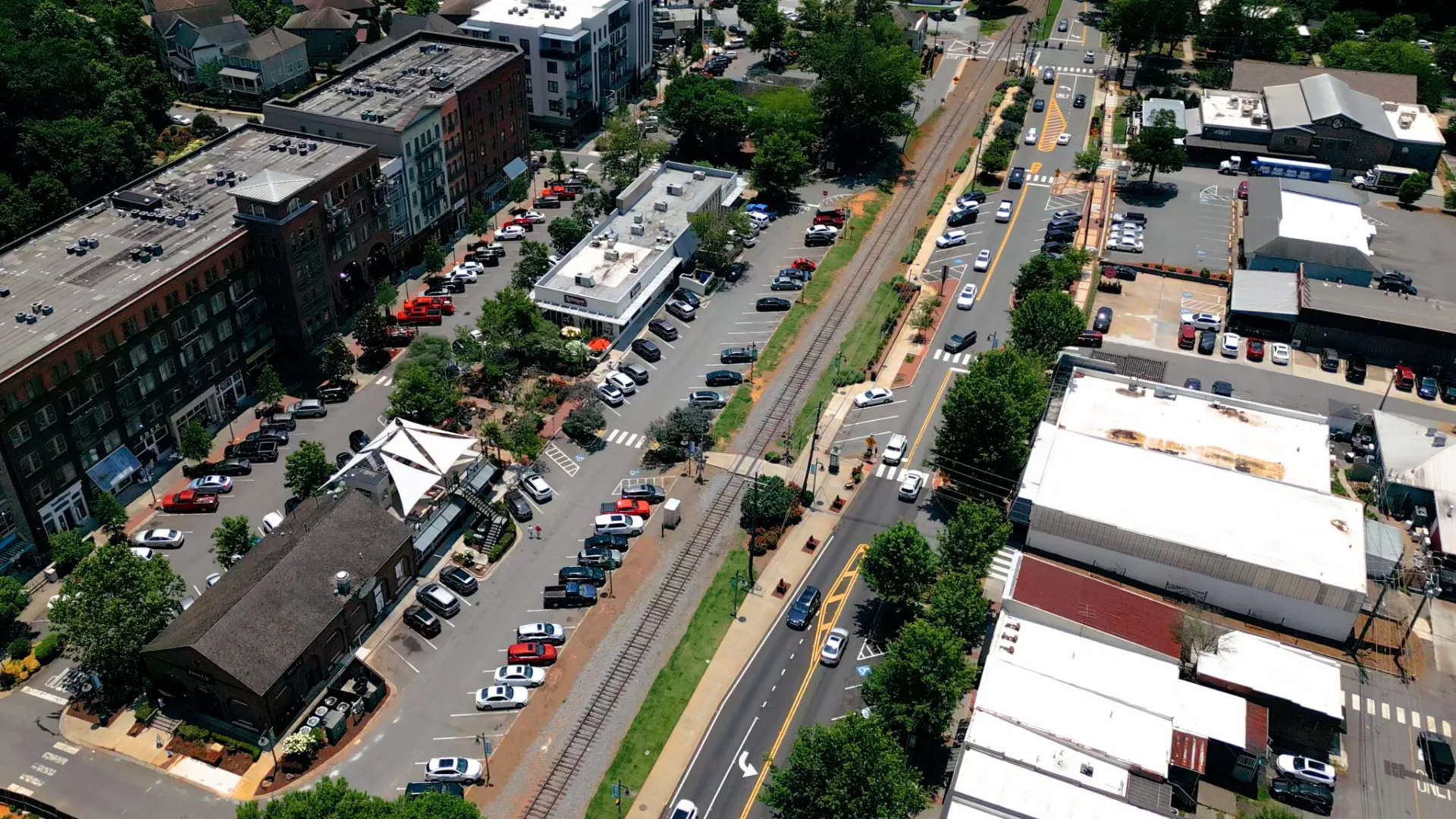 Aerial view of a busy urban street with parked cars, buildings, trees, and a train track running through the center.