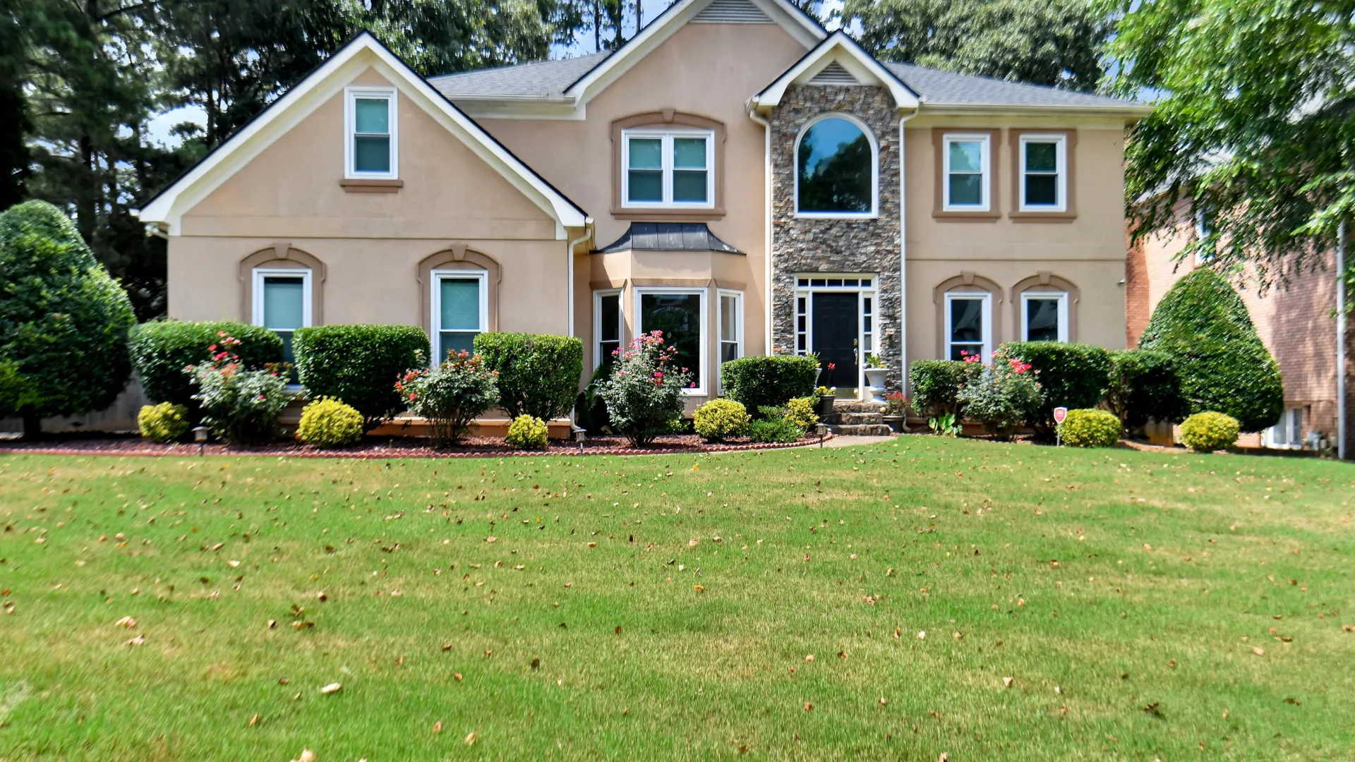 Large beige two-story house in Snellville, GA with stone accents, manicured lawn, shrubs, and tall trees in the background