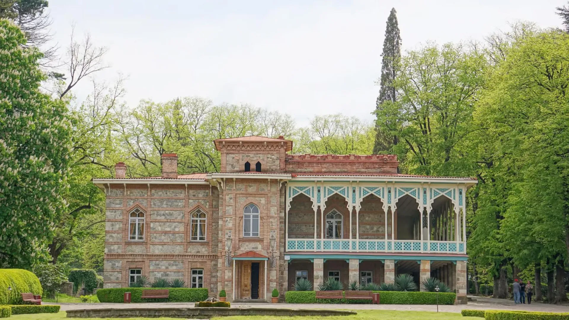 Historic brick mansion with ornate balconies surrounded by lush green trees and manicured gardens on a clear day