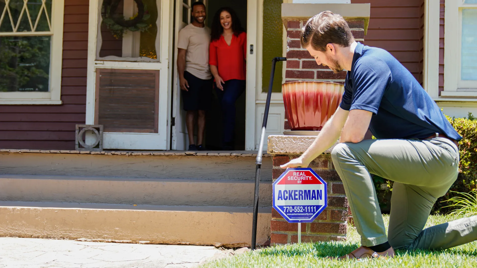 Man installing Ackerman security sign in front yard while couple watches from front porch of home.