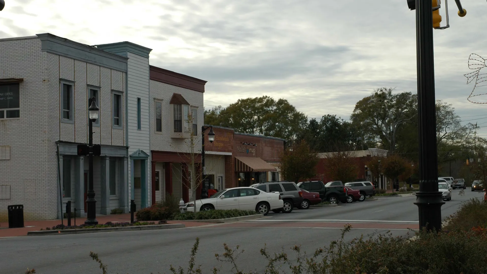 Small town street with parked cars, brick buildings, street lamps, and overcast sky in late afternoon.