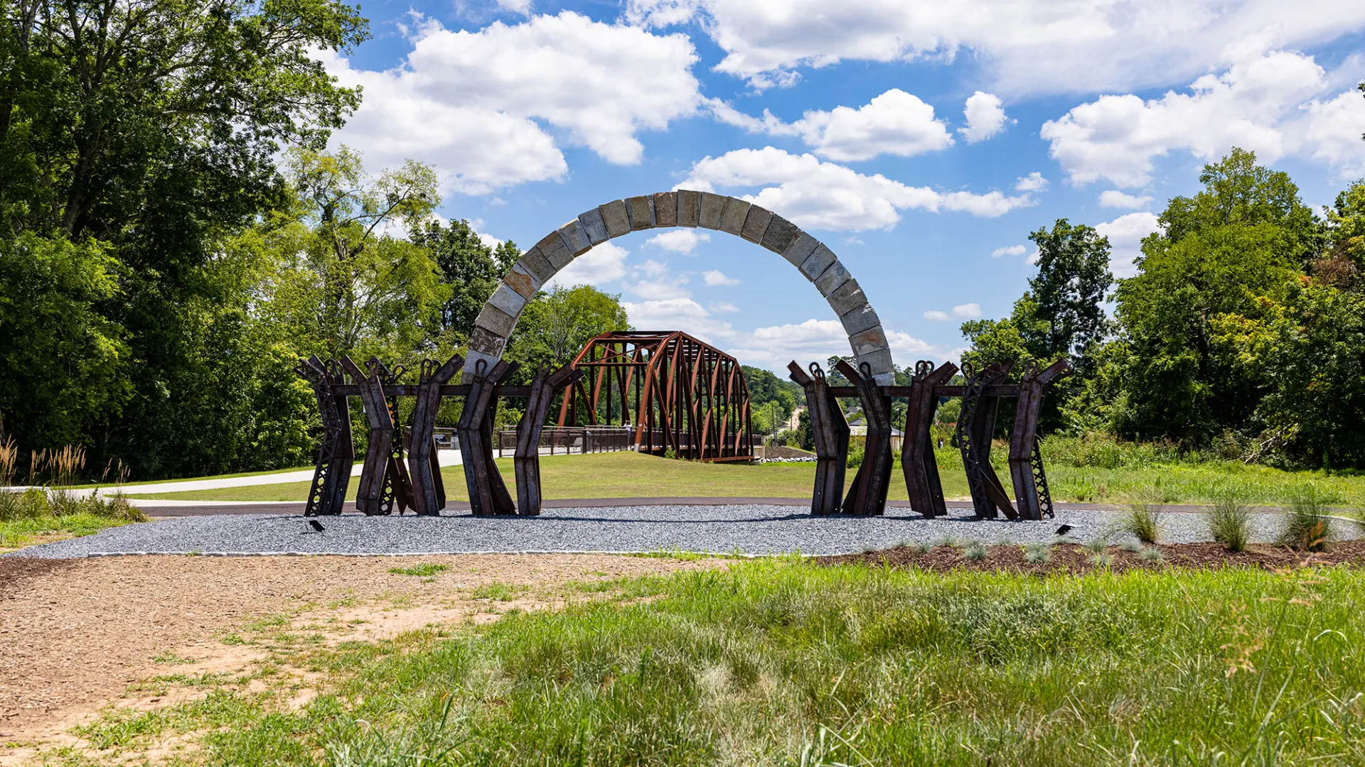 Stone arch and metal sculptures frame a rusted metal bridge with greenery and blue sky background