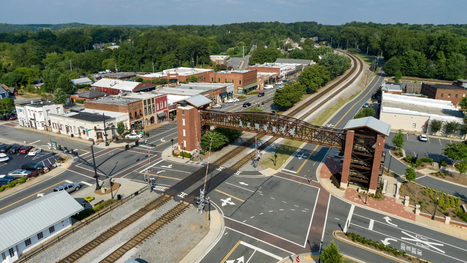 Aerial view of a small town railroad crossing with a pedestrian bridge and downtown buildings surrounded by trees.