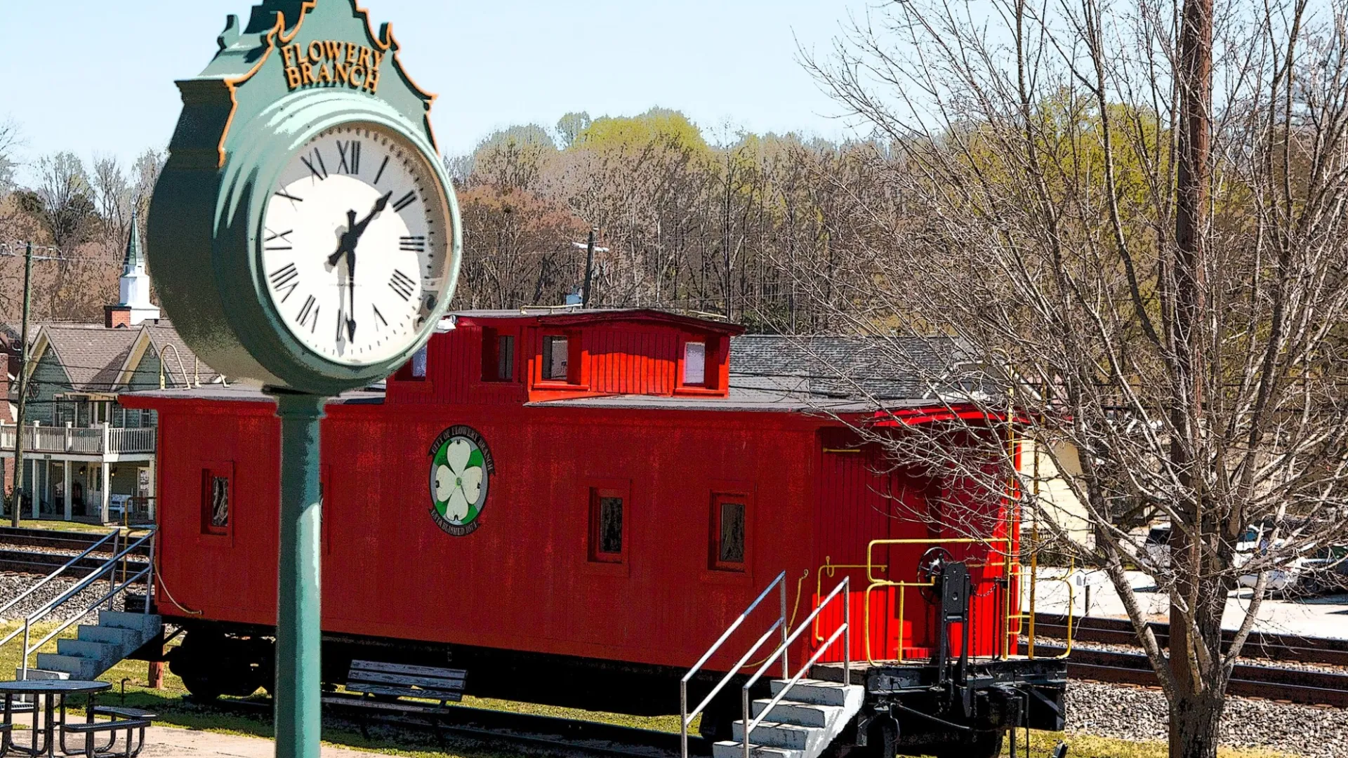 Historic red train car near a Flowery Branch clock in a small town park setting with bare trees and evergreen background.