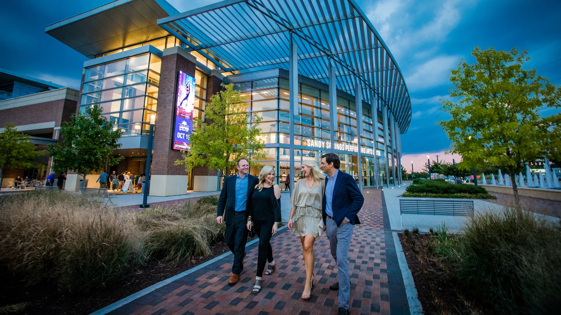 Four people walking and chatting outside a modern performing arts center during evening with trees and fountains.