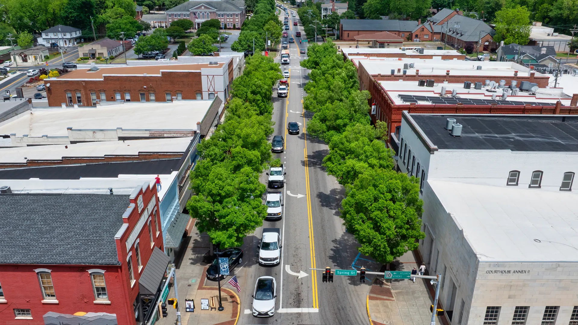 Aerial view of a tree-lined street with parked cars and buildings on both sides in a small town downtown area.
