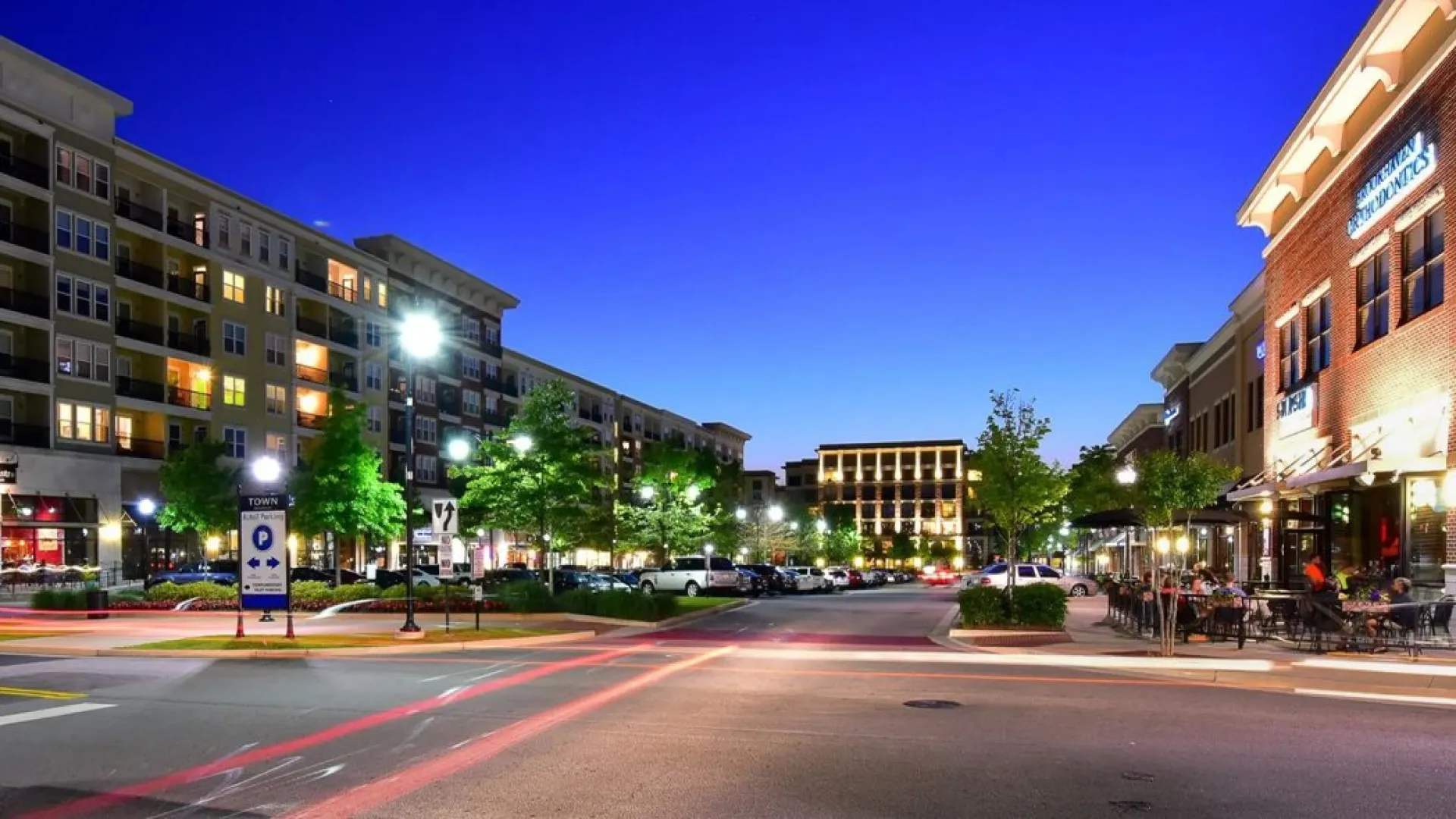 Night view of an urban street with modern apartments, lit trees, parked cars, and outdoor dining on the right.