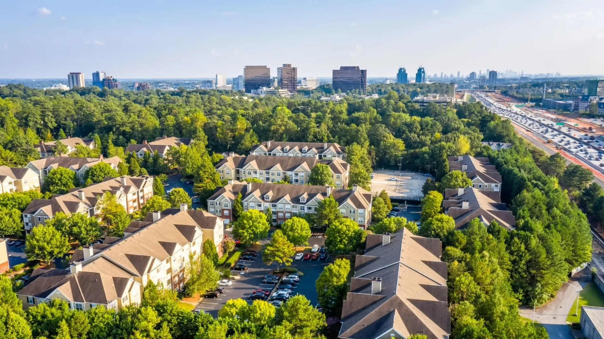 Aerial view of suburban apartment complex surrounded by trees with city skyline and highway in background.
