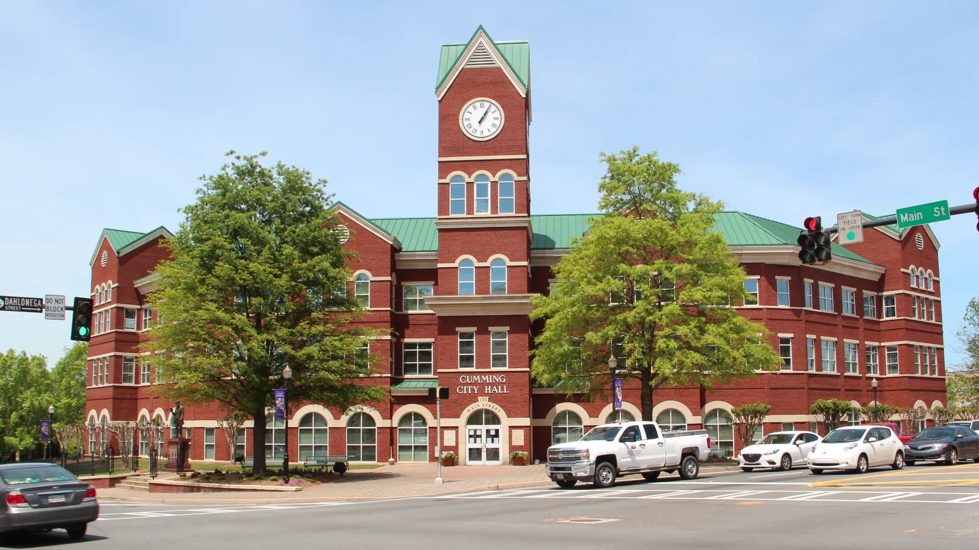 Brick city hall building with clock tower, surrounded by greenery and parked vehicles.