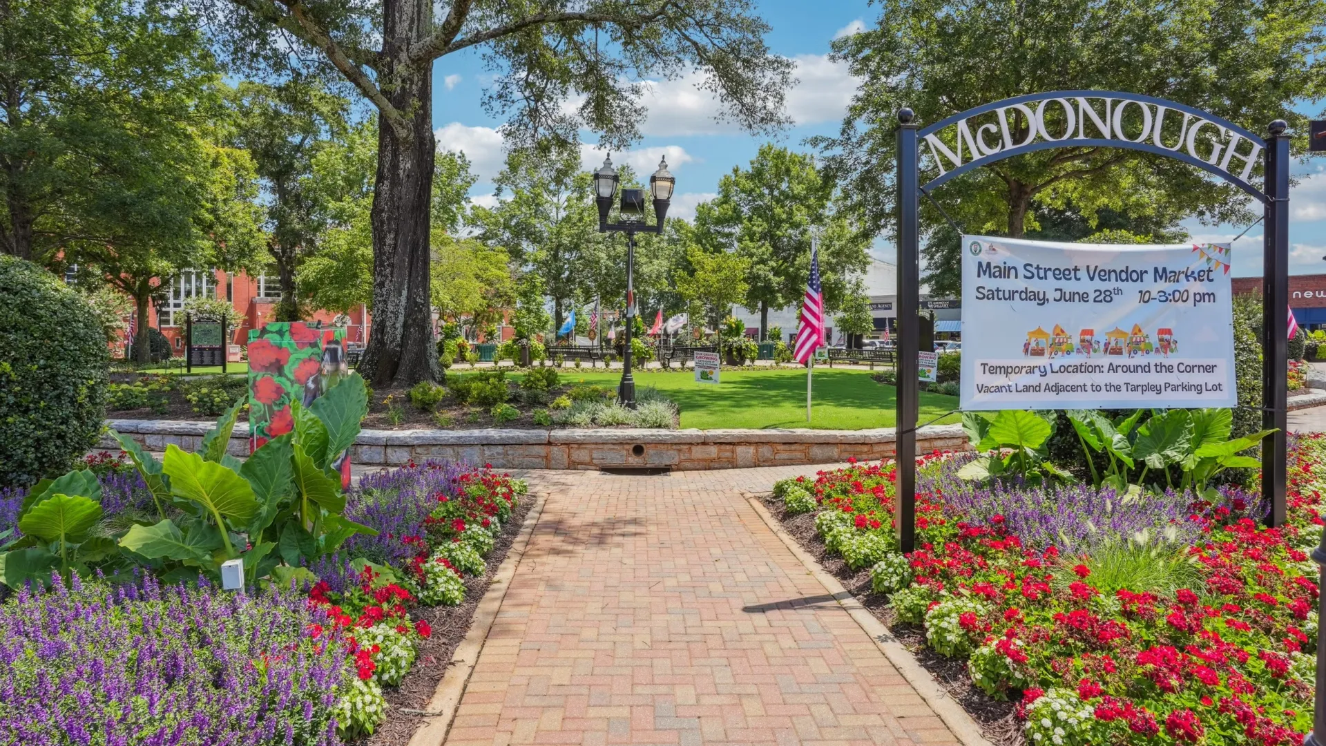 Brick pathway through vibrant flower beds and greenery under a sunny blue sky in McDonough park.