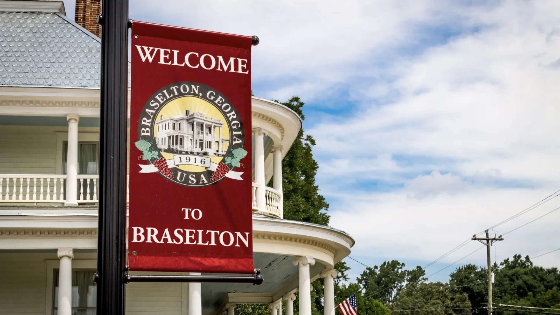 Red welcome sign for Braselton, Georgia hanging from lamppost in front of historic white building with columns.