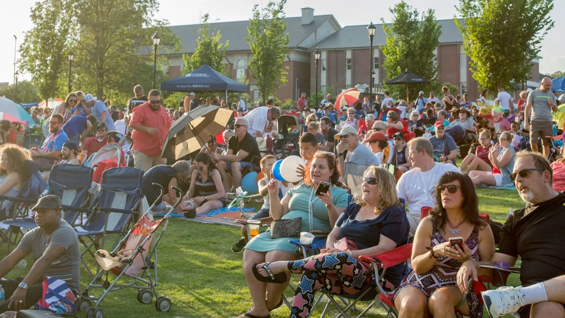 Large diverse crowd gathered outdoors on a sunny day, sitting on chairs and blankets enjoying an event on grassy lawn.