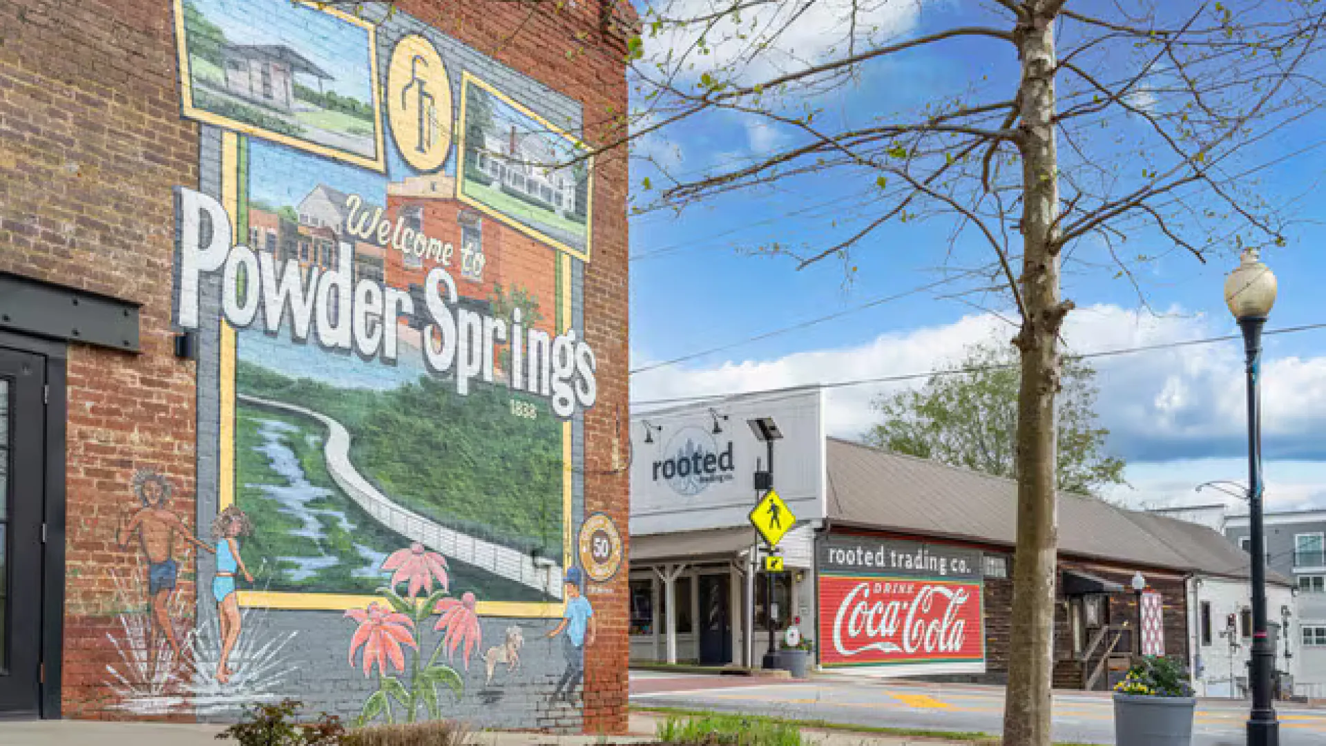 Mural welcoming visitors to Powder Springs on a brick wall by a street with Coca-Cola signage and tree in foreground.