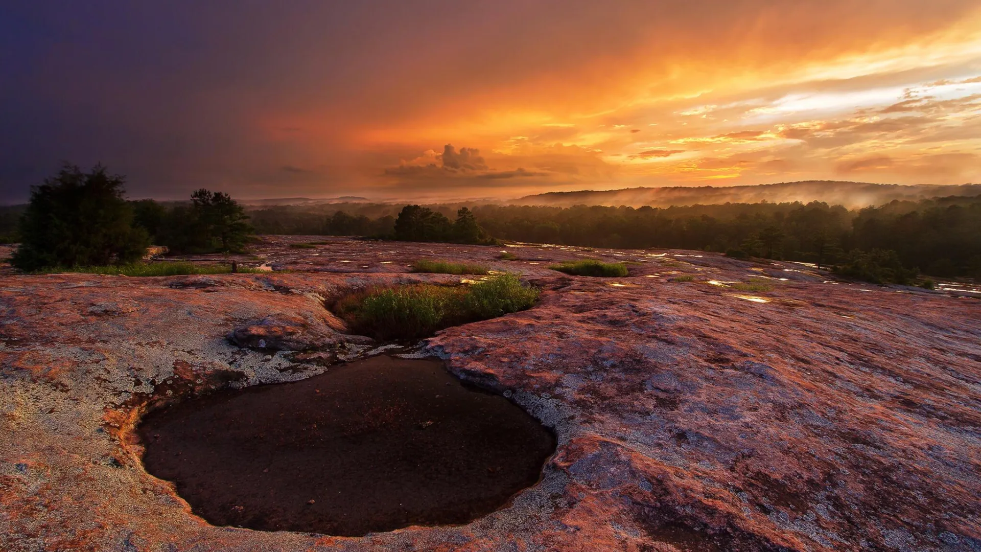 Sunset over rocky terrain with shallow pools and misty forest in the background under colorful sky.