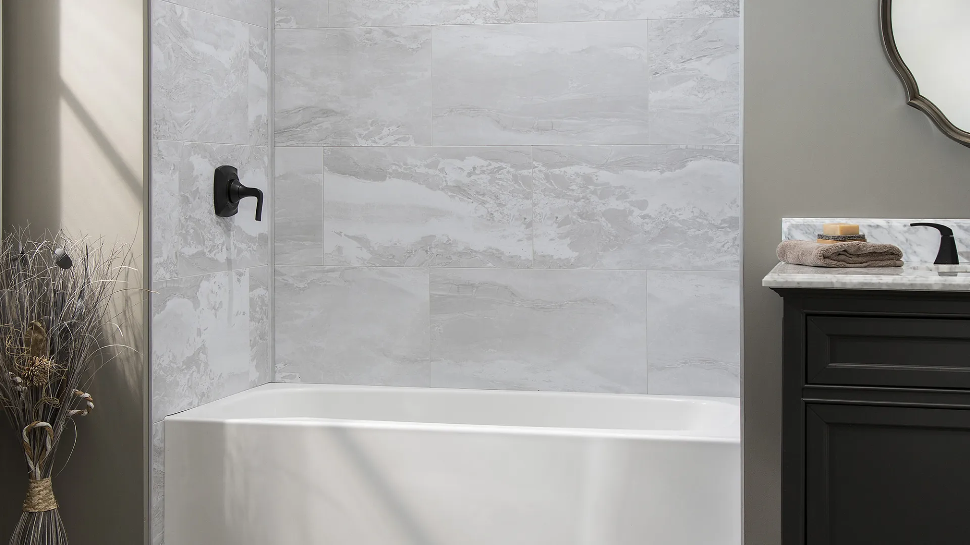 Modern bathroom with white bathtub, gray marble tile shower, black fixtures, and dark vanity with mirror.