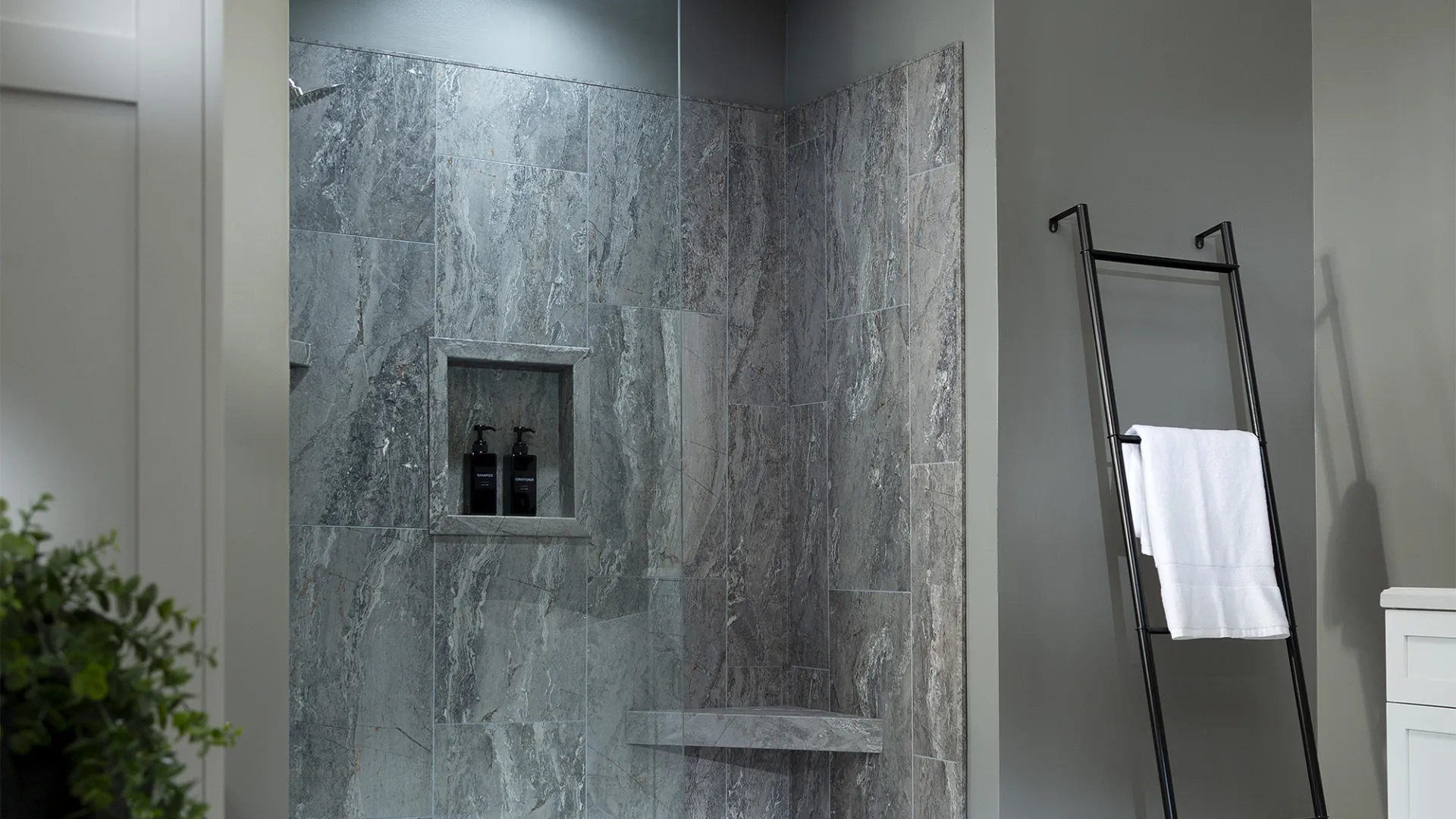 Modern bathroom with gray stone tiled walk-in shower, glass partition, black ladder towel rack, and white cabinetry.