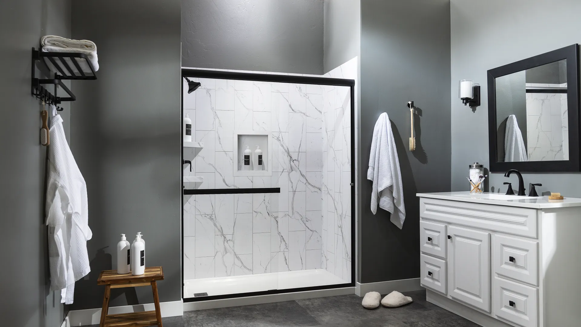 Modern bathroom with gray walls, white vanity, glass shower with marble tiles, and wooden stool with toiletries.