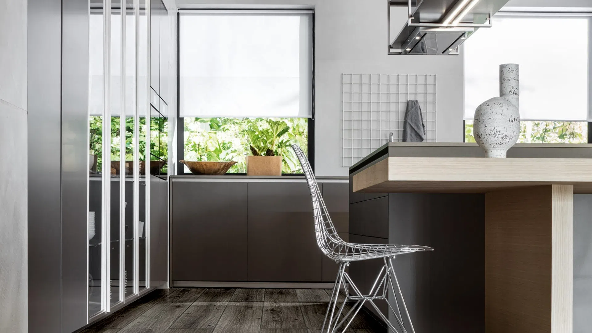 Modern kitchen with dark wood flooring, grey cabinets, a wireframe chair, and hanging shelves with kitchenware.