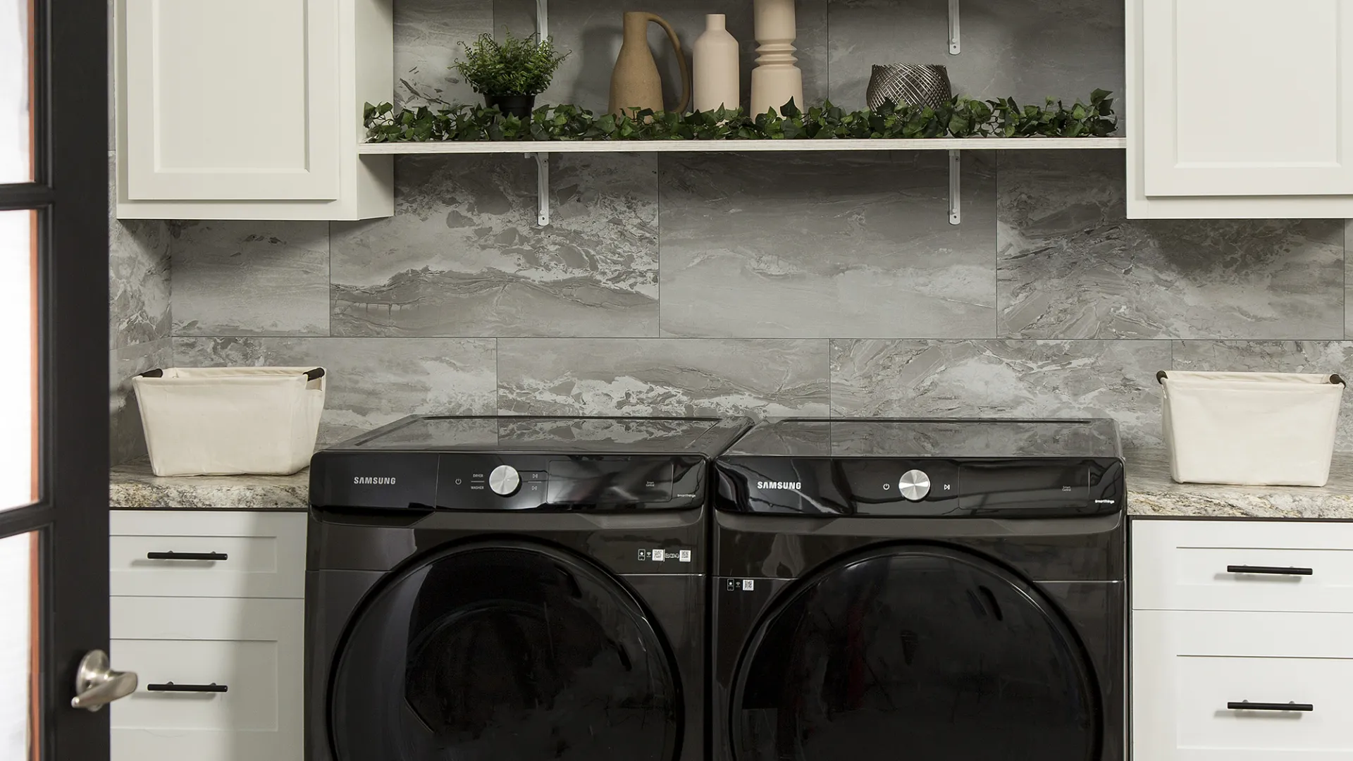 Modern laundry room with black Samsung washer and dryer, white cabinets, marble wall, and decorative shelves.