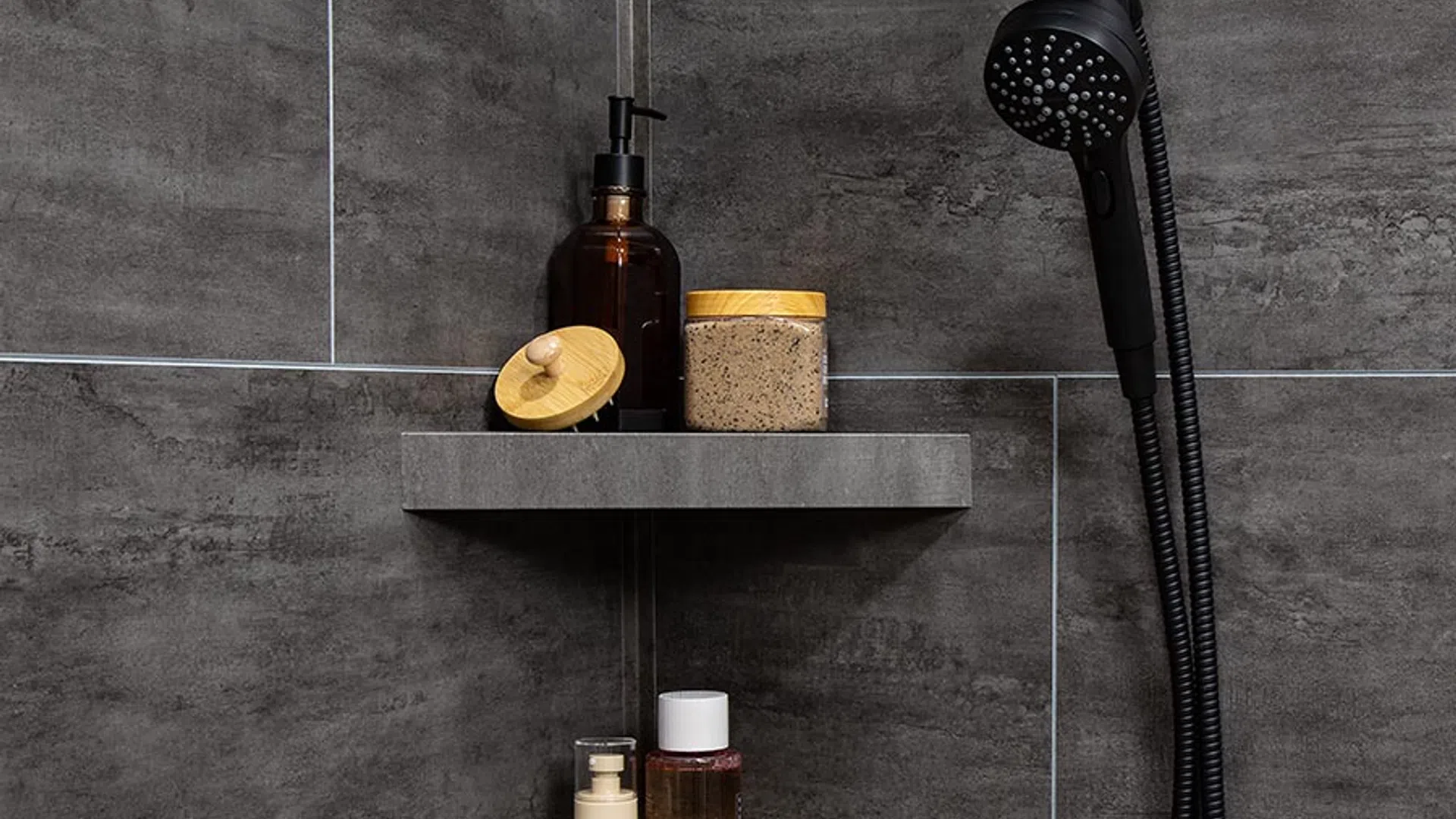 Modern shower corner with dark gray tiles, built-in shelves holding bottles and scrub, and a black handheld showerhead.