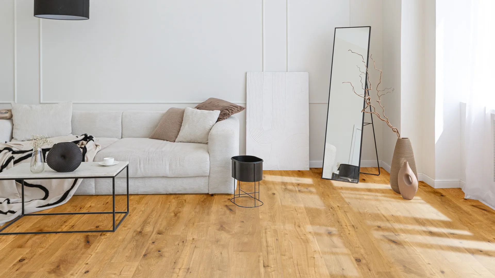 Bright modern living room with white sofa, wooden floor, black lamp, tall mirror, and decorative vases by window.