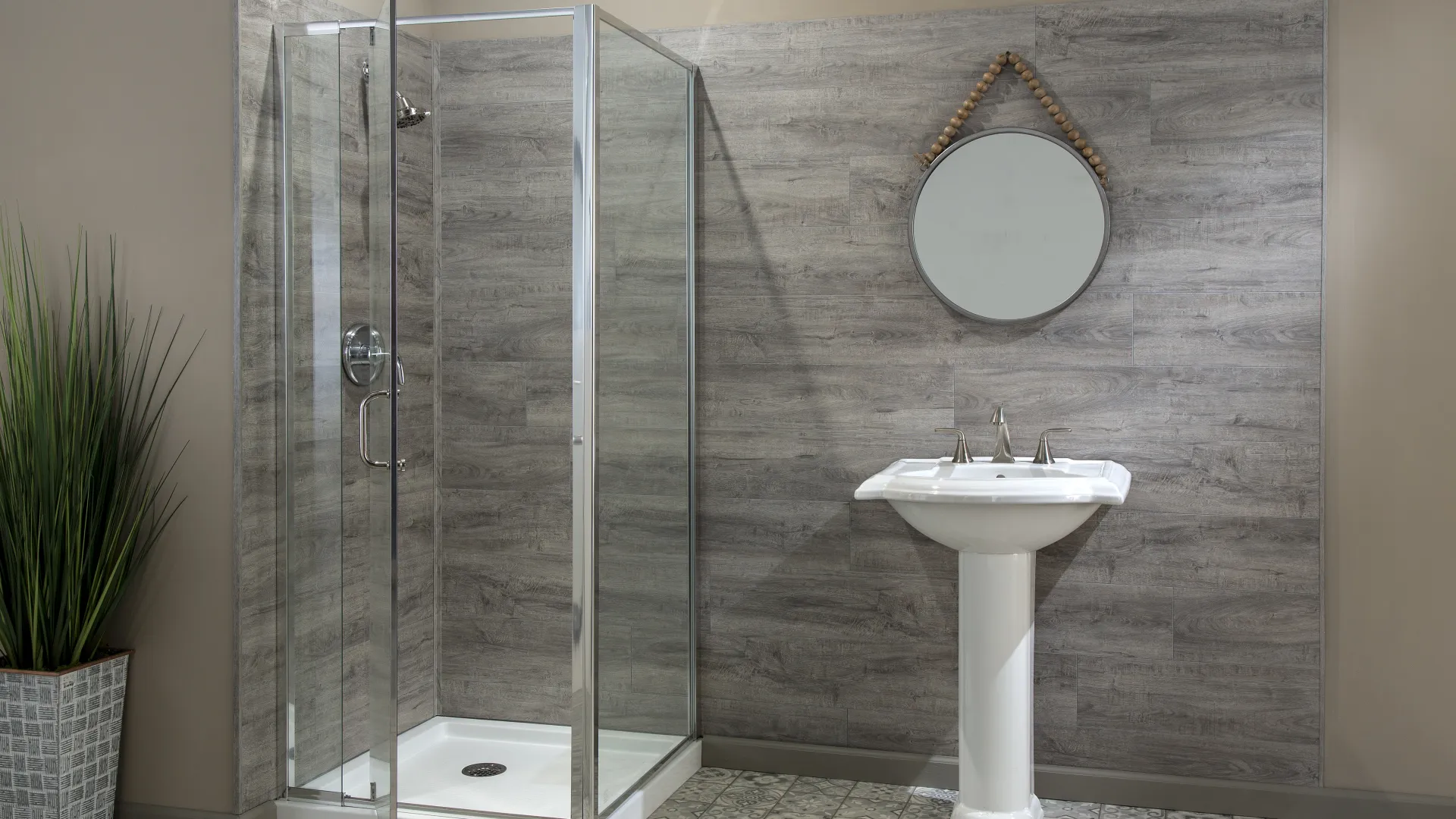 Modern bathroom with glass shower enclosure, pedestal sink, round mirror, and potted tall plant.