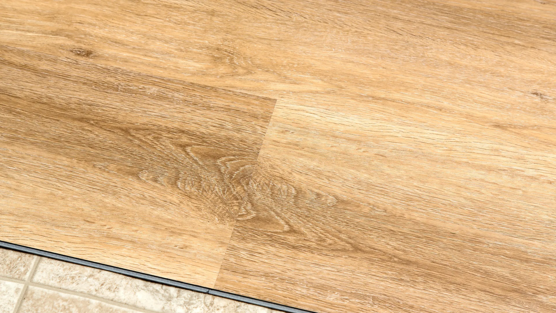 Close-up of a wood grain laminate floor panel next to beige ceramic tile with grout lines.